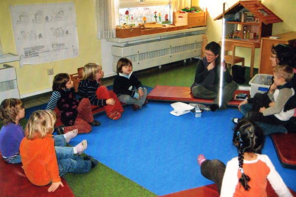 Eine Kindergruppe sitzt kreisförmig mit der blinden Besucherin zusammen auf dem Boden. Die Kinder bestaunen den Blindenlangstock von der blinden Frau

